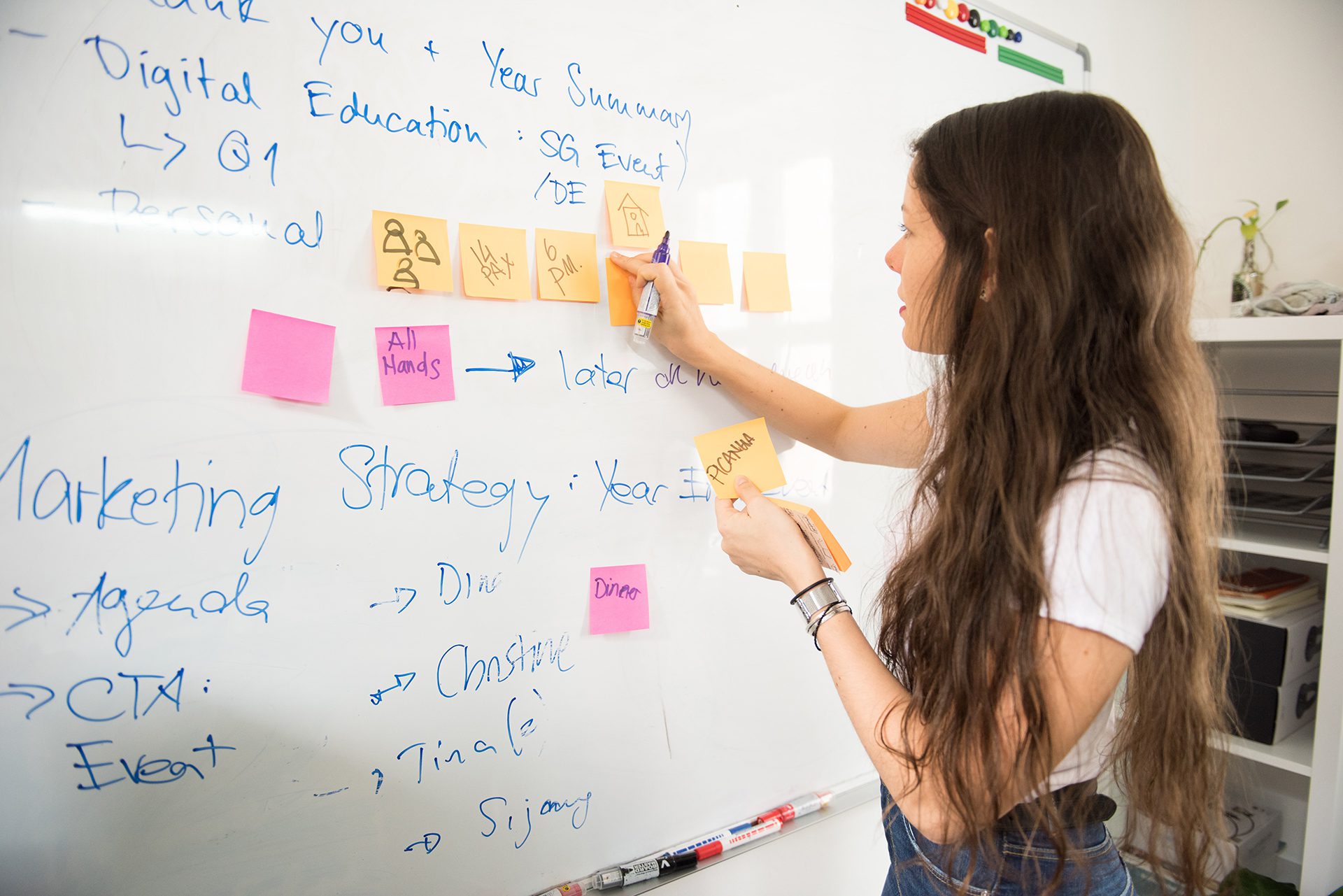 Woman with long brown hair writing on post it notes stuck to a whiteboard filled with notes