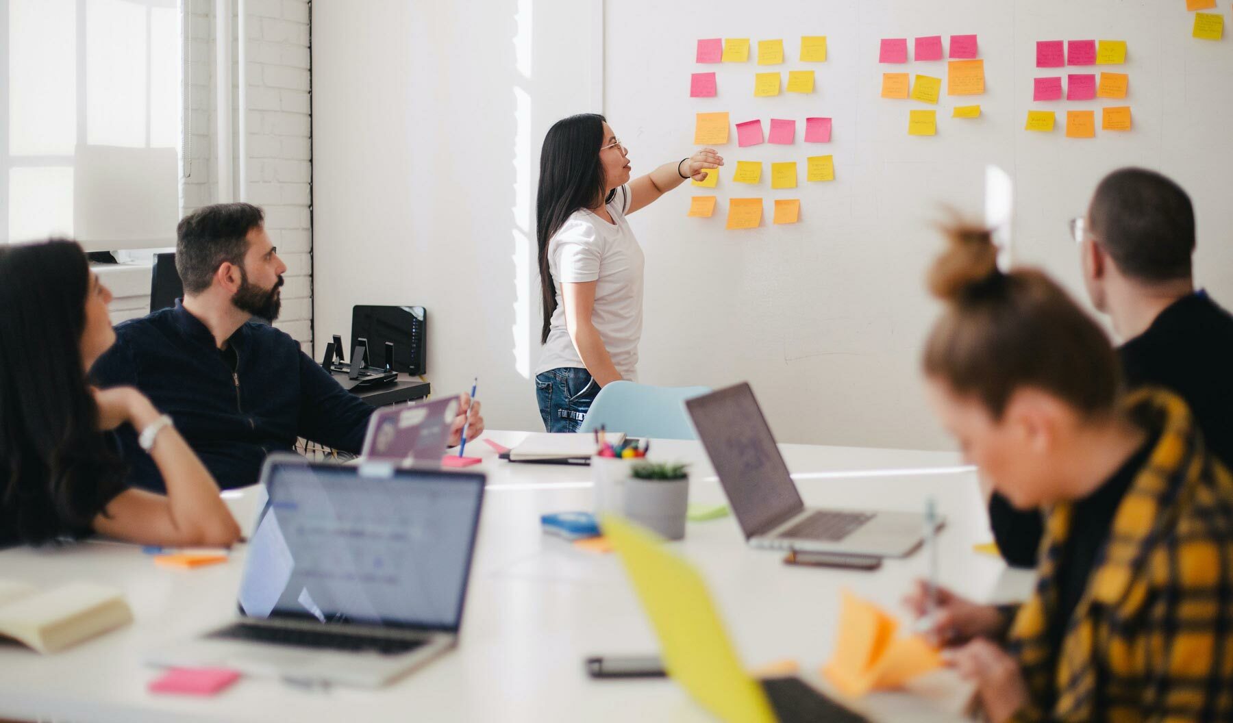 People sat at desks with laptops looking at person at whiteboard pointing to sticky notes